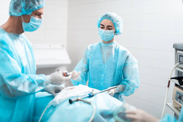 Young nurse in protective mask giving surgical instrument to surgeon - Foto, Bild