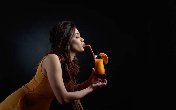 junge schöne Mädchen in einem orangefarbenen Kleid mit perfektem Make-up hält ein Glas Cocktail. Cocktail Tequila Sonnenaufgang garniert mit Kirsche und Orangenscheiben. Kopierraum. - Foto, Bild