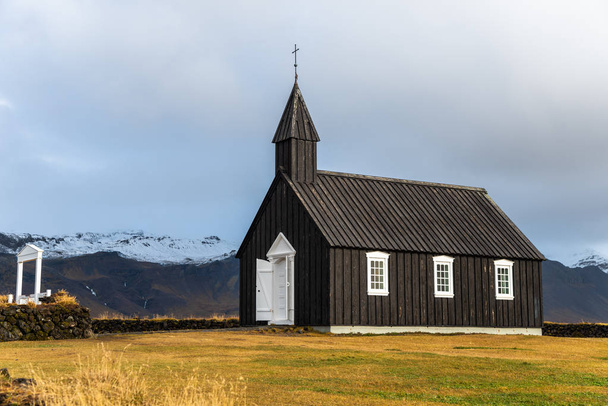 Petite église en bois noir avec des montagnes enneigées en arrière-plan dans l'ouest de l'Islande par un jour d'automne pluvieux
 - Photo, image