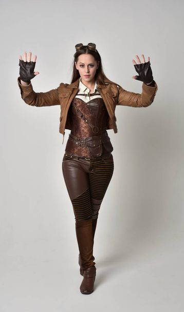 retrato de longitud completa de chica morena con traje steampunk de cuero marrón. pose de pie sobre fondo gris estudio
. - Foto, Imagen