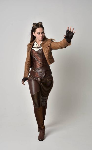 full length portrait of brunette  girl wearing brown leather steampunk outfit. standing pose on grey studio background. - Foto, afbeelding