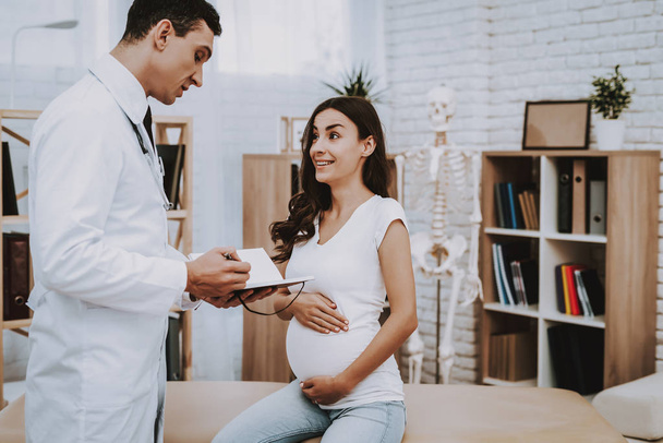 Pregnant Girl at the Gynecologist Doctor. Doctor is Young Man. Doctor is Writing Information About a Patient. Girl is Sitting on Couch and Touching Her Belly. Girl is Smiles. People Located at Clinic. - Zdjęcie, obraz