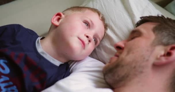 Little boy cuddling up with his dad in bed. They're talking about something interesting before they go to sleep. - Video
