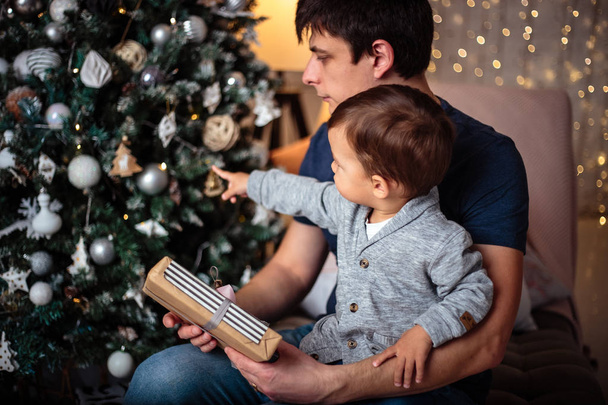 Küçük oğlu rahat bir ceket ve kot babasıyla birlikte Noel ağacının yanında oturuyor. Genç adam ve oğlu - Fotoğraf, Görsel