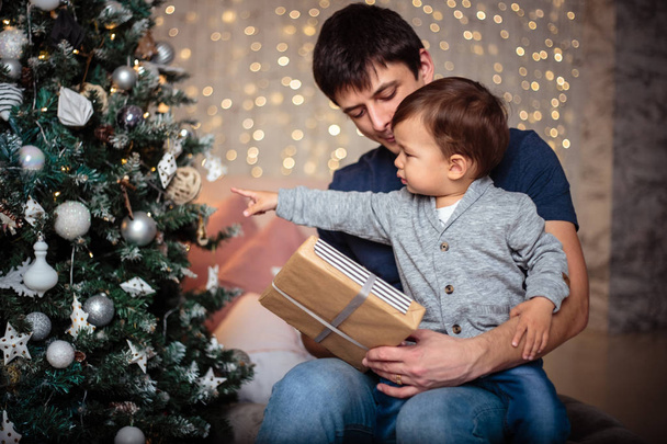 Little baby boy in a casual jacket and jeans sitting near christmas tree with his dad. Young man and his son - Фото, изображение