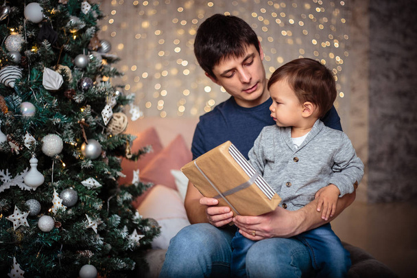 Little baby boy in a casual jacket and jeans sitting near christmas tree with his dad. Young man and his son - Фото, изображение