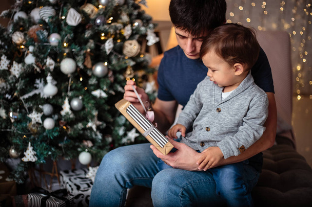 Little baby boy in a casual jacket and jeans sitting near christmas tree with his dad. Young man and his son - Φωτογραφία, εικόνα