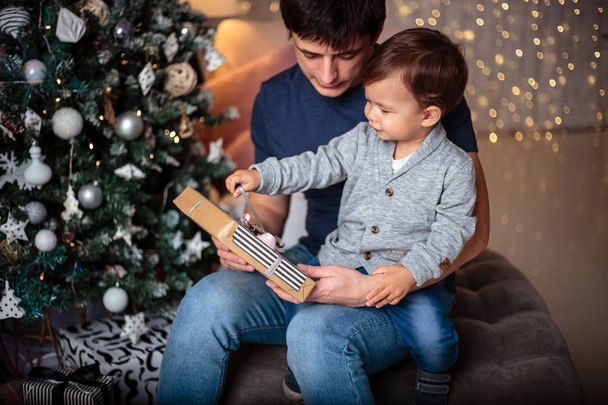 Little baby boy in a casual jacket and jeans sitting near christmas tree with his dad. Young man and his son - Fotoğraf, Görsel