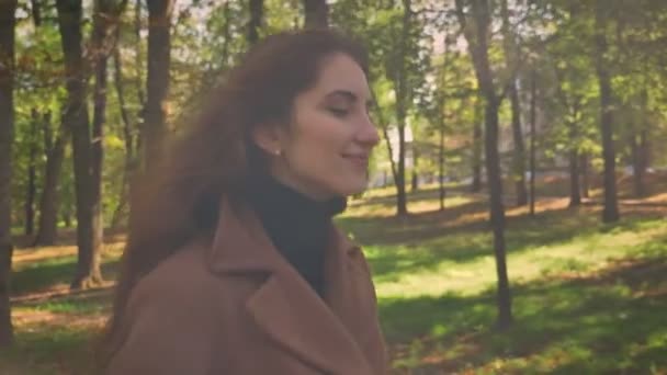 Shooting of smiling beautiful female walking towards camera with her brunette wavy hair on the wind and sunny park on background, moving model illustration - Filmati, video