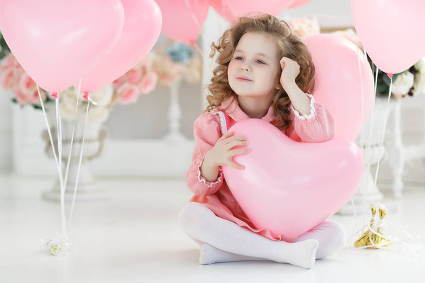 Valentine's Day, postcard. Small girl with blond curly hair poses in a white studio surrounded by pink balloons. A beautiful preschool girl in a white studio with heart-shaped pink balloons. Valentine's Day, love, birthday. - Zdjęcie, obraz