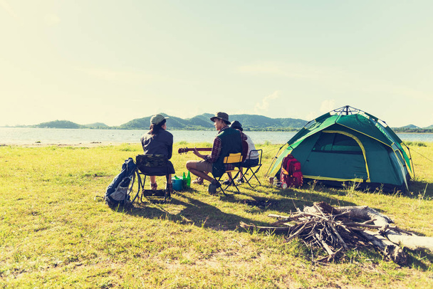 Group of travelers camping and doing picnic and playing music together. Mountain and lake background. People and lifestyle. Outdoors activity and leisure theme. Backpacker and Hiker. Back view angle - Photo, Image