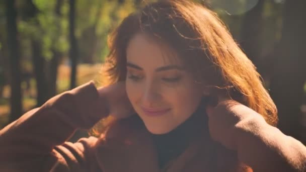 Close up caucasian woman is strengthening her brown hair and smiling calmly glancing at camera, green trees on background - Imágenes, Vídeo