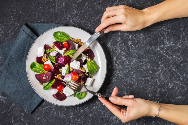dish with a salad of feta, beetroot, cherry tomatoes and walnuts on a stone background. Female hands hold fork and knife. Top view - Photo, image