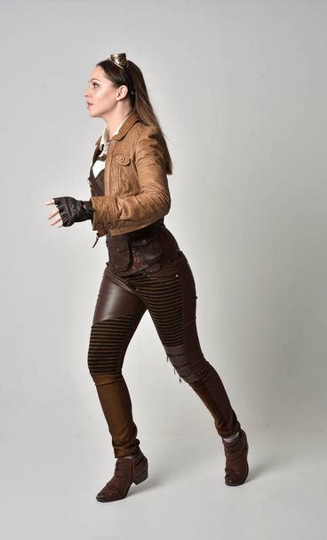 full length portrait of brunette  girl wearing brown leather steampunk outfit. standing pose on grey studio background. - Fotografie, Obrázek
