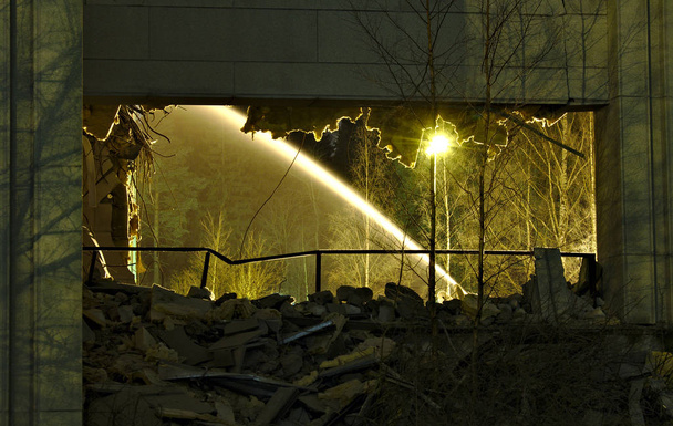 Widok przez zawalony budynek, tylko ściana wciąż stoi. Woda z uszkodzonej rury wodnej tworzącej fontannę oświetloną światłami ulicznymi. - Zdjęcie, obraz