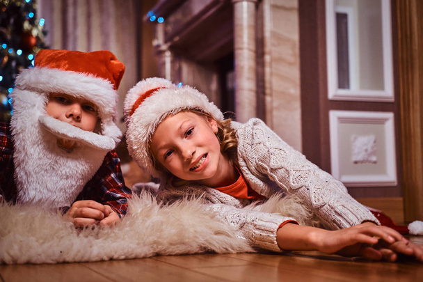 Gelukkig broer en zus dragen KERSTMUTS liggend op een tapijt van bont in de buurt van een kerstboom thuis. - Foto, afbeelding