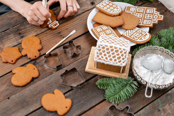 若い女の子は、冬の朝雪だるまクリスマスの形のジンジャー クッキーを飾る。女性は、はちみつジンジャーブレッド クッキーにアイシングを描画します。木製の茶色のテーブル。コピー スペース. - 写真・画像