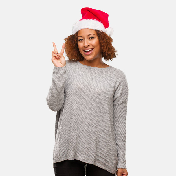 Fiatal fekete nő, fárasztó egy santa kalap, szórakoztató és vidám csinál egy gesztus, a győzelmet - Fotó, kép