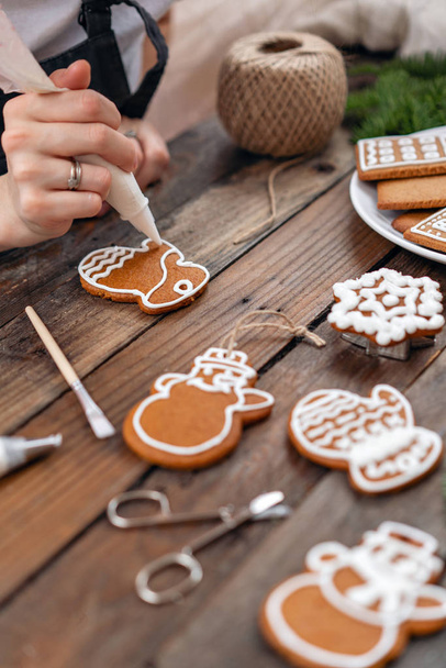 Cookies in Form von Fäustlingen. Ein junges Mädchen dekoriert Ingwer-Plätzchen an Heiligabend. Frau zeichnet Sahnehäubchen auf Honig-Lebkuchen. Holztisch braun. Kopierraum. - Foto, Bild