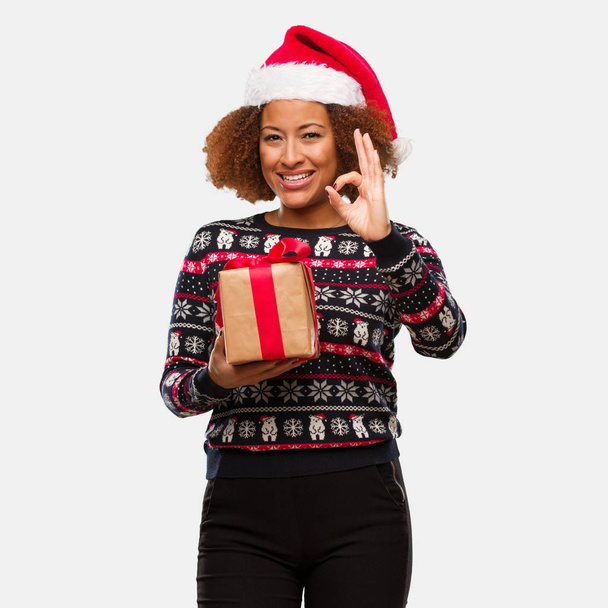 Νεαρή μαύρη γυναίκα που κρατά ένα δώρο για την ημέρα των Χριστουγέννων, χαρούμενα και με αυτοπεποίθηση κάνει εντάξει χειρονομία - Φωτογραφία, εικόνα