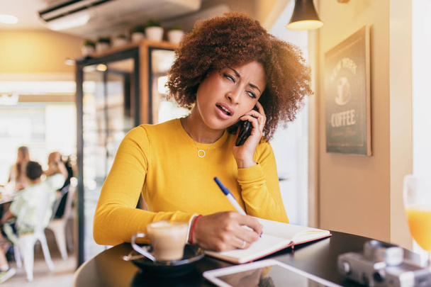 Jeune femme afro au café, parlant sur téléphone portable, très stressée et dépassée, écrivant dans un carnet
. - Photo, image