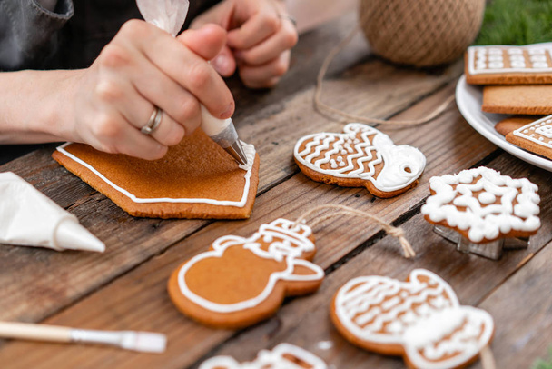 若い女の子は、冬の朝ジンジャー クッキー クリスマスを飾る。女性は、はちみつジンジャーブレッド家にアイシングを描画します。木製の茶色のテーブル。領域をコピーします。空白ビスケット ジンジャーブレッドの家を飾るために準備ができて. - 写真・画像