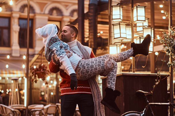 Mladý romantický pár těší trávit čas spolu o Vánocích na ulici zdobí krásná světla. Šťastný mladý kluk drží své dívky v náručí a líbání. - Fotografie, Obrázek