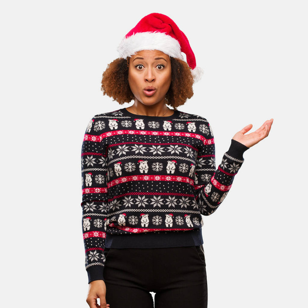 Mujer negra joven en un suéter de Navidad de moda con estampado sosteniendo algo en la palma de la mano
 - Foto, imagen