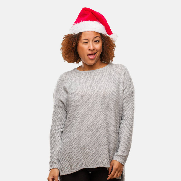 Young black woman wearing a santa hat winking, funny, friendly and carefree gesture - Photo, Image