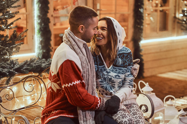 Una joven pareja romántica que viste ropa de abrigo al aire libre en Navidad, sentada en un banco en la calle de la noche decorada con hermosas luces
. - Foto, Imagen