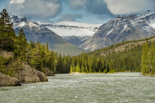 BANFF, AB, CANADÁ - JUNIO 2018: Vista del paisaje del río Bow, que fluye a través de Banff, con altas montañas cubiertas de nieve en el fondo
. - Foto, imagen
