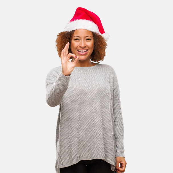 jovem preto mulher vestindo um santa chapéu alegre e confiante fazendo ok gesto
 - Foto, Imagem