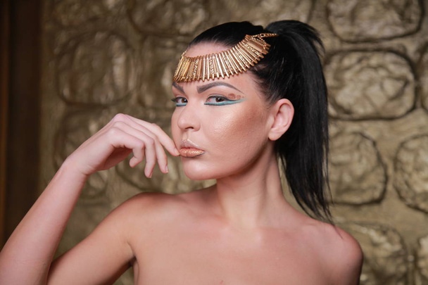 ziemlich gebräunte Brünette posiert. Reiche Luxusfrau im ägyptischen Stil mit Schmuck auf goldenem Hintergrund. - Foto, Bild