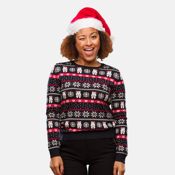 Νεαρή μαύρη γυναίκα σε ένα καθιερώνον τη μόδα πουλόβερ Χριστουγέννων με εκτύπωσης winking, αστείο, φιλικό και ξένοιαστη κίνηση - Φωτογραφία, εικόνα
