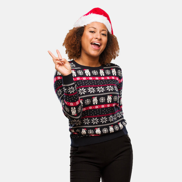 Fiatal fekete nő egy trendi karácsonyi pulóver nyomtatása, csinál egy gesztus, a győzelmet - Fotó, kép