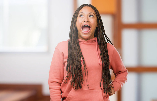 Retrato de uma jovem negra usando tranças gritando com raiva, expressão de loucura e instabilidade mental, boca aberta e olhos meio abertos, conceito de loucura
 - Foto, Imagem