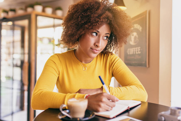 Młoda kobieta afro w kawiarni, ona myśli o pomysł aby napisać. - Zdjęcie, obraz