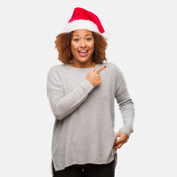 Jovem negra vestindo um chapéu de Papai Noel sorrindo e apontando para o lado
 - Foto, Imagem