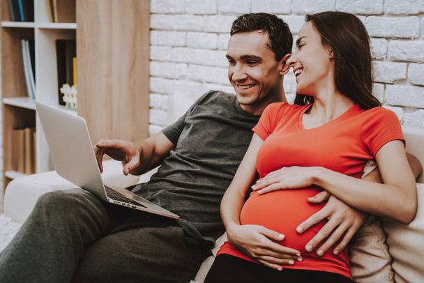 Couple is Watching Video on Laptop. Couple is a Man and Pregnant Woman. Girl is Touching Her Belly. Persons is Smiling. Husband is Hugging His Wife. People is Sitting on Couch. People Located at Home. - Zdjęcie, obraz