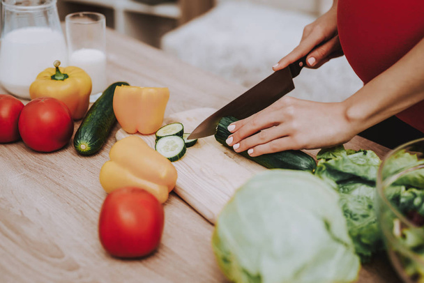 Woman is Cooking Salad from Fresh Vegetables. Woman is a Young Pregnant Girl. Girl is Cutting the Cucumber a Knife. Vegetables, Milk and Salad in Bowl on Table. Girl Located at Home. Table Closeup. - Photo, Image
