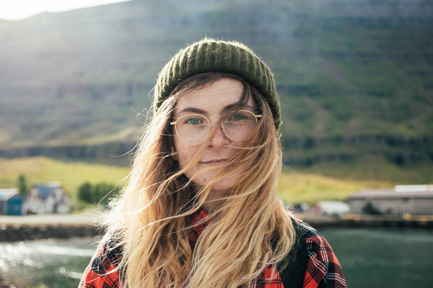 Portrait of young woman with blonde hair and green hat standing outdoor in nature with windy weather  - Photo, Image