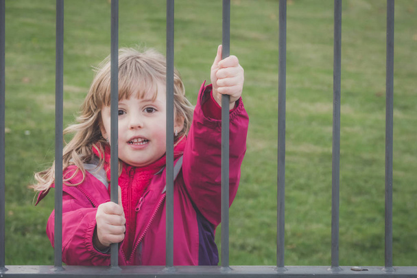Little cute Caucasian girl dressed in pink jacket standing behind metal gates leading to the playground - Foto, Bild