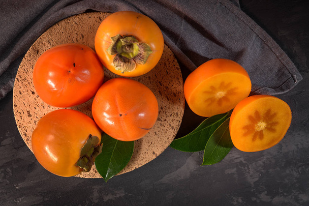 Ripe persimmon fruits in a cork plate on dark kitchen counter top. - Foto, immagini