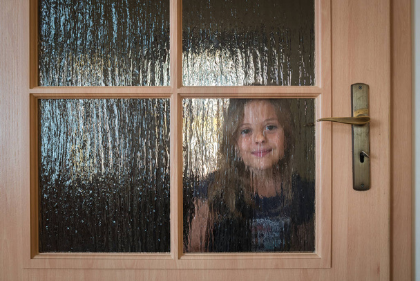 Portré egy aranyos kis kaukázusi lány bujkál az ajtó mögött üveg ablakok játék közben bújócska - Fotó, kép