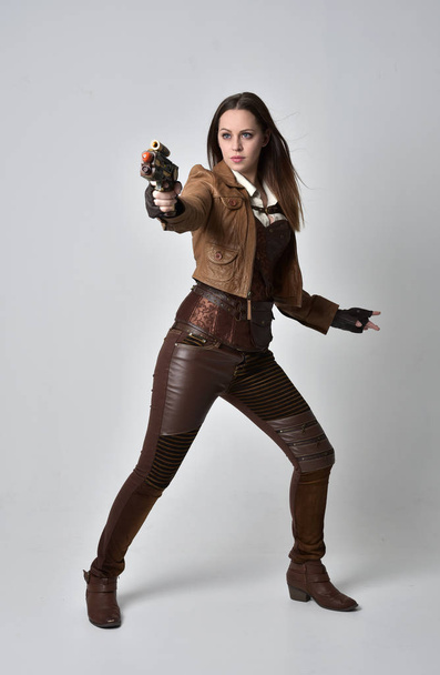 Ganzkörperporträt eines brünetten Mädchens im braunen Leder-Steampunk-Outfit. Stehende Pose mit Pistole, auf grauem Studiohintergrund. - Foto, Bild