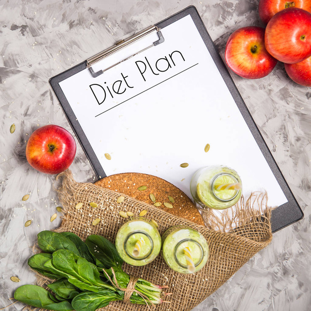 Het dieetplan is geschreven op een wit vel op de tablet onder de flessen van groene smoothies, spinazie en appels op een grijze tafel met jute. Gewichtsconcept verlies. Bovenaanzicht - Foto, afbeelding
