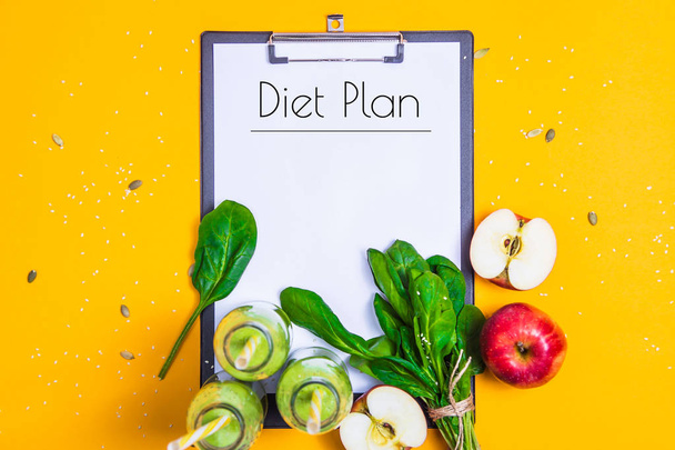 The diet plan is written on a white sheet on the tablet among the bottles of green smoothies, spinach and apples on a bright yellow background. Slimming and detox concept. Top view - Fotoğraf, Görsel