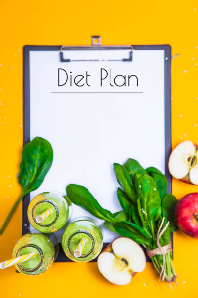 The diet plan is written on a white sheet on the tablet among the bottles of green smoothies, spinach and apples on a bright yellow background. Slimming and detox concept. Top view - Foto, immagini