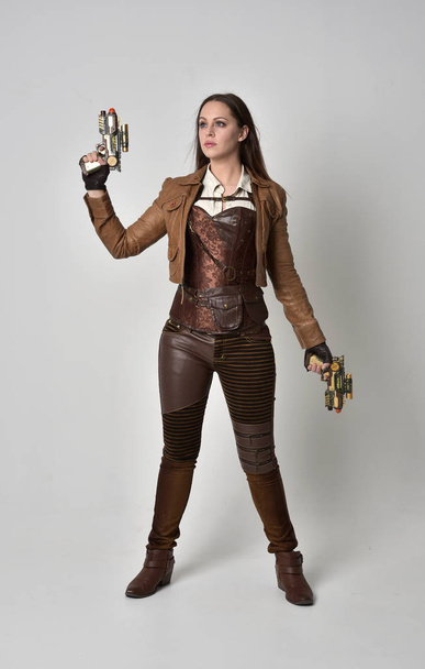 full length portrait of brunette  girl wearing brown leather steampunk outfit. standing pose holding a gun, on grey studio background. - Zdjęcie, obraz