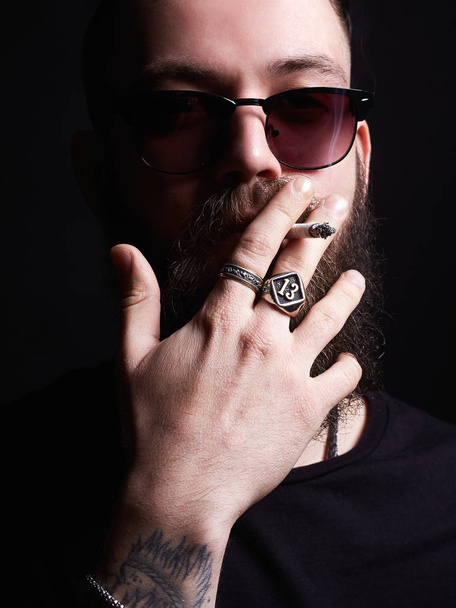 przystojny brutalny Brodaty palacza w okulary przeciwsłoneczne. Hipster tattoed chłopiec z papierosa i pierścienie na rękę - Zdjęcie, obraz